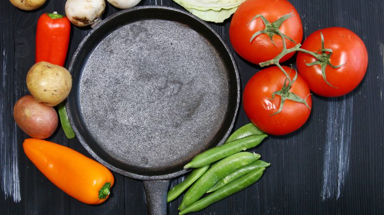 Top view of a cast iron skillet surrounded by vegetables like tomatoes, potatoes, peas, peppers, cabbage and mushrooms