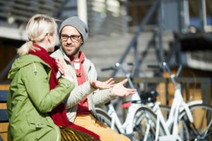 Young Caucasian bearded man wearing glasses chatting with girlfriend while sitting on bench near parked bicycles on sunny autumn day