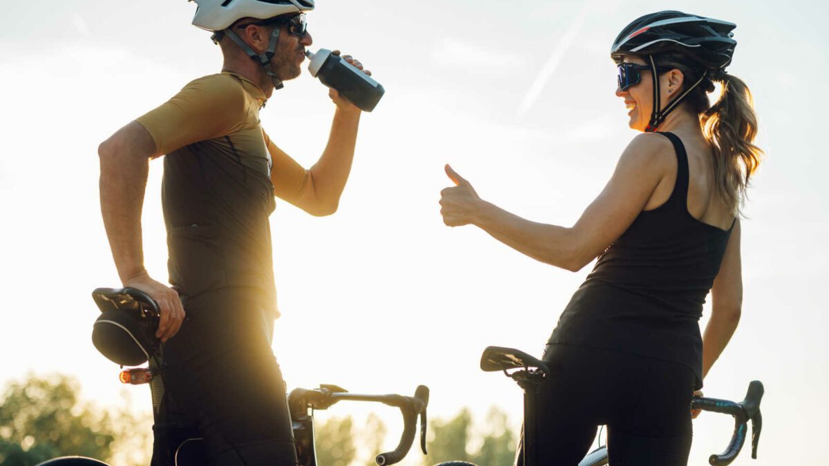 Professional riders cycling on racing bikes in the nature. Active lifestyle and sport concept. Sporty couple of cyclist riding a bicycle on the road against the sunset. Focus on a man drinking water.