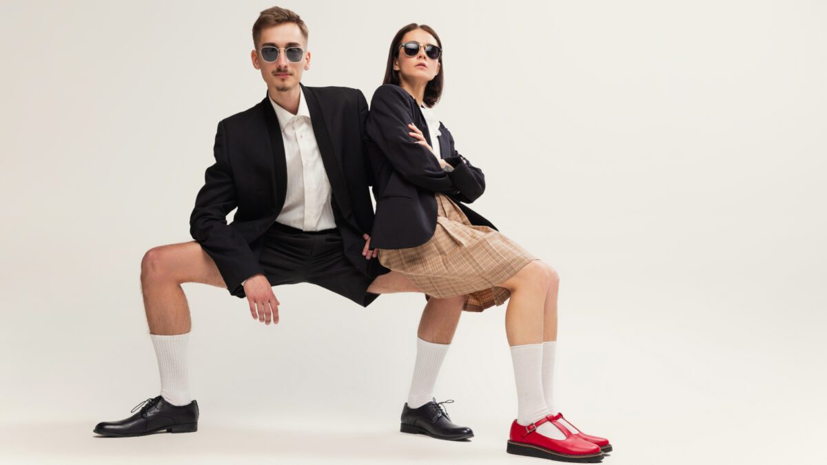 Full-length portrait of stylish young man and woman in retro suit posing isolated over grey studio background. Sitting on knee. Concept of retro fashion, art photography, style, queer, beauty